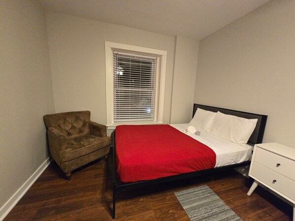 Une chambre avec un lit simple, une table de chevet, une chaise, une fenêtre avec des stores et un plancher en bois.