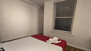 Hotellihuone, jossa on yksi sänky, punainen sängynpäällinen, ikkuna, jossa on sälekaihtimet, puulattia ja kattovalaisin.