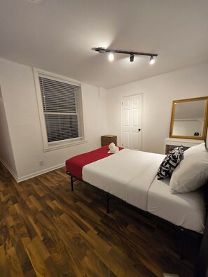Une chambre à coucher avec un lit, une fenêtre avec des stores, une porte, une table de chevet et un miroir.
