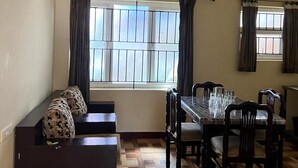A dining area with a table and chairs, a sofa with patterned cushions, and a window with curtains. - Essar Cottage (Ootacamund)