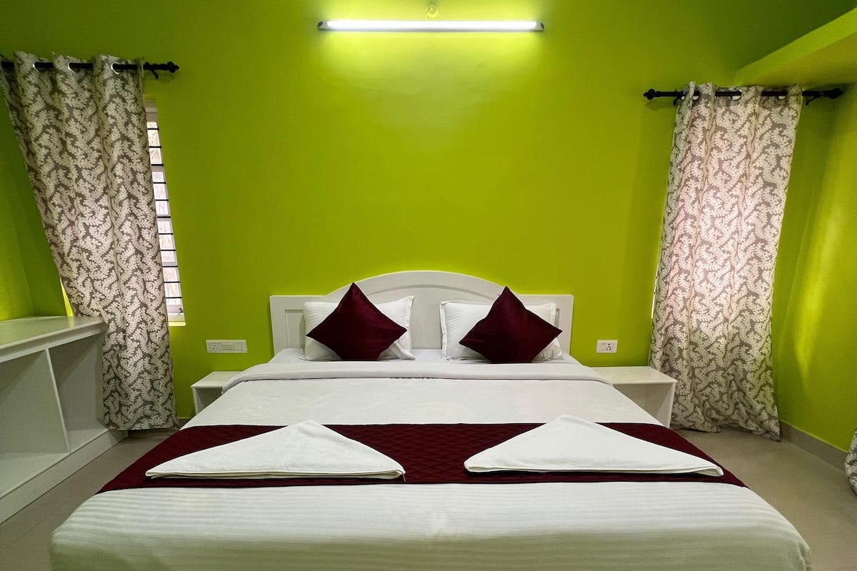 A neatly made bed with white linens and two red pillows, a white headboard, and a green wall with a ceiling light fixture.