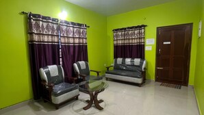 A living room with green walls, two sofas, a glass coffee table, and a wooden door. - Essar Cottage (Ootacamund)