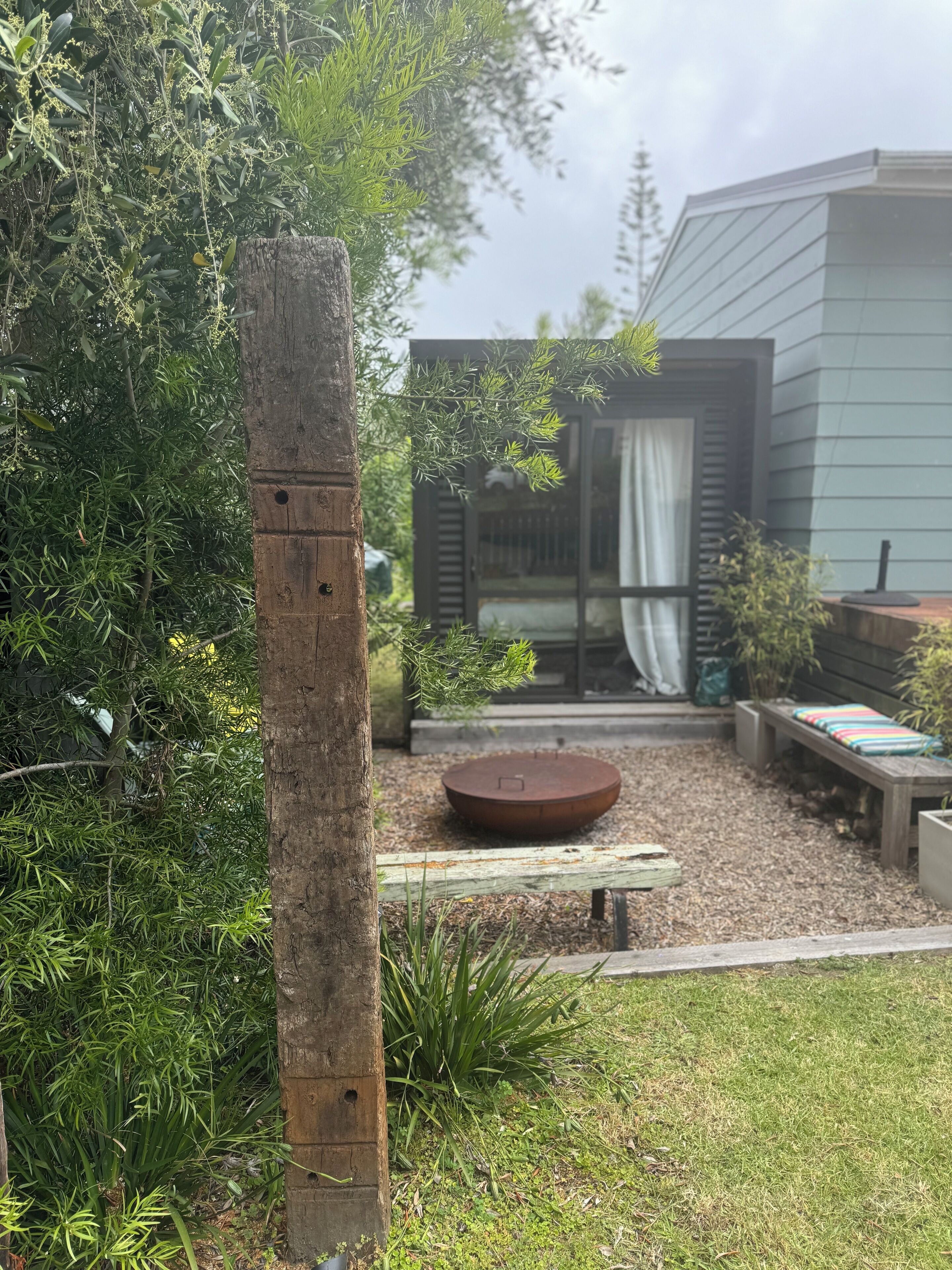 A garden with a wooden post, a round planter, and a bench. There is a building with a glass door and a curtain in the background.