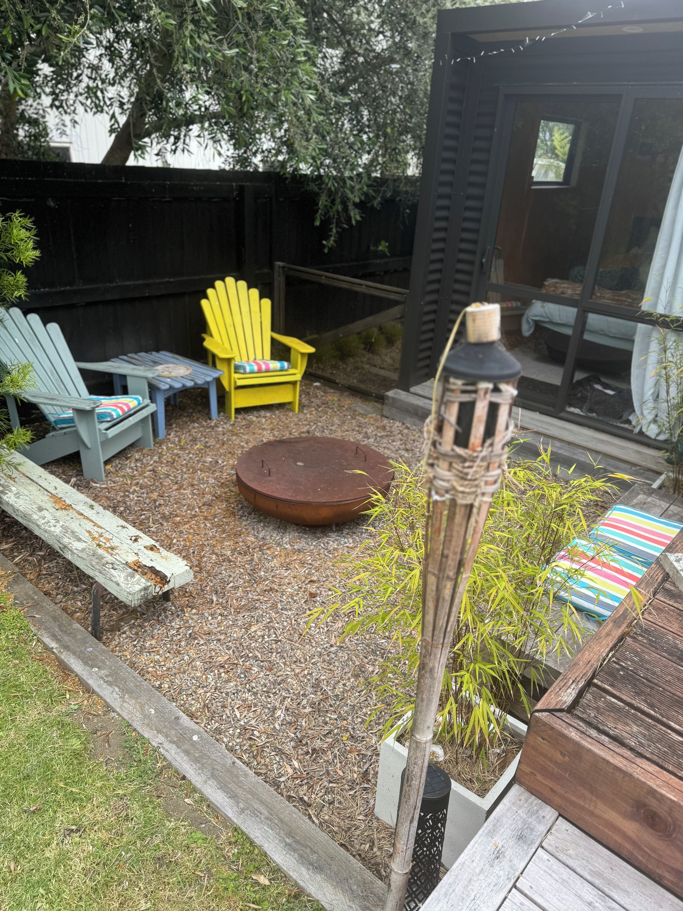 A backyard with a wooden deck, a round table, two Adirondack chairs, a small garden with bamboo, and a metal lantern.