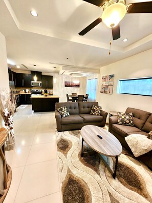 A modern living room with a sofa set, a coffee table, and a TV. There is a kitchen in the background with dark cabinets and a microwave. - Fully Furnished 3BR/2BA Condo. Close to Airport, Hospitals, Shopping & Dinning (McAllen)