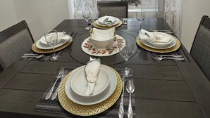 A dining room with a dark wooden table set for four, featuring gold-rimmed plates and clear glassware. A chandelier hangs from the ceiling, and the room has large windows with sheer curtains. - Fully Furnished 3BR/2BA Condo with Balcony. Close to Airport, Shopping & Dinning (McAllen)