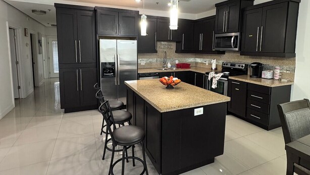 A modern kitchen with dark cabinetry, a central island with bar stools, a microwave, and a refrigerator. - Fully Furnished 3BR/2BA Condo with Balcony. Close to Airport, Shopping & Dinning (McAllen)