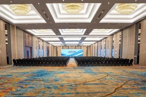 Ein großer Konferenzraum mit Reihen von Stühlen, die auf eine Bühne zeigen, einem gemusterten Teppich und einer Kristallleuchter.