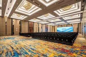 Ein großer Konferenzraum mit Teppichboden, Reihen von Stühlen, einer Bühne mit Bildschirm und einem modernen Deckenstil.