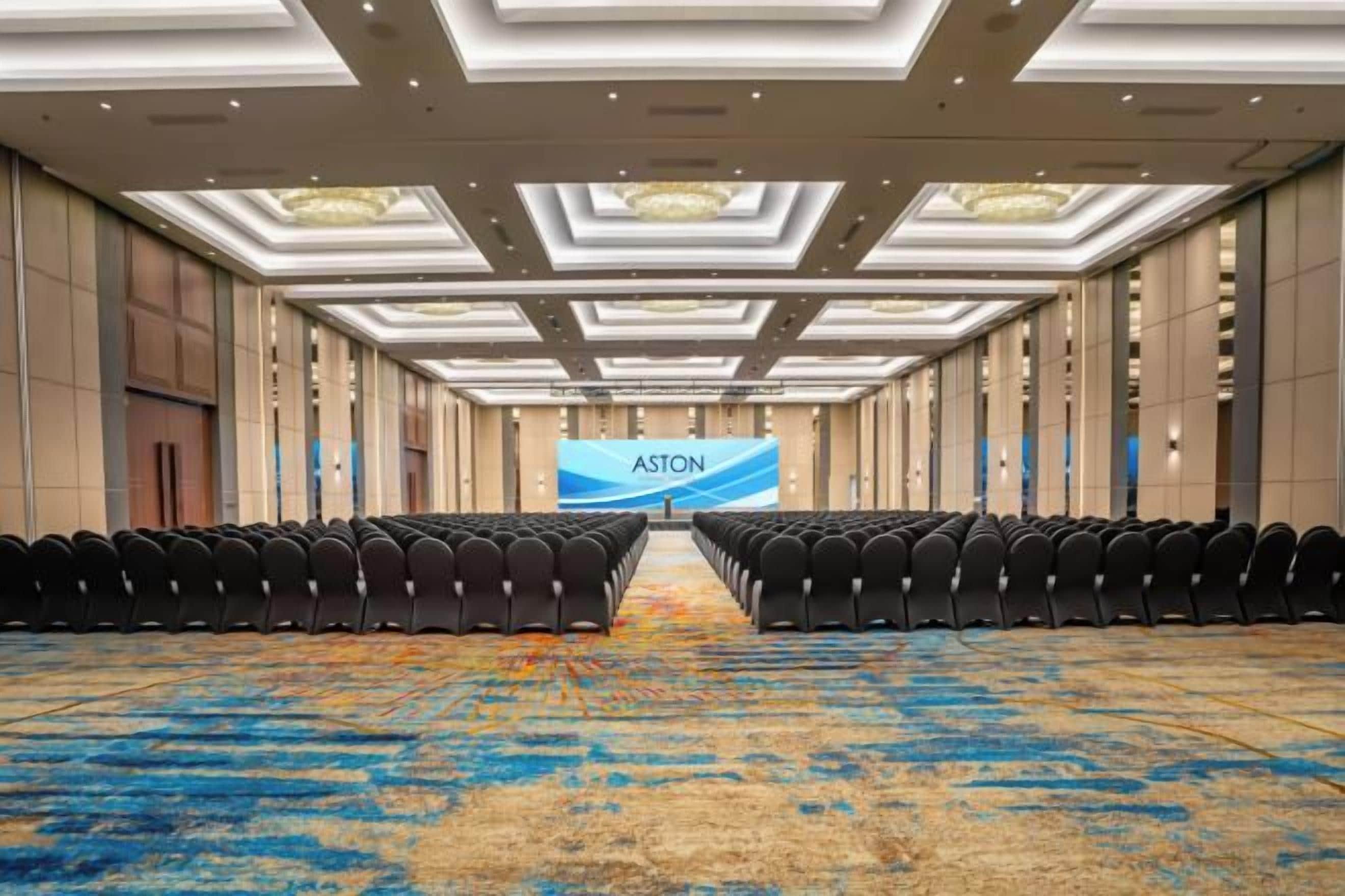 Una sala conferenze con file di sedie, un grande schermo che mostra 'ASTON' e un tappeto a motivi.
