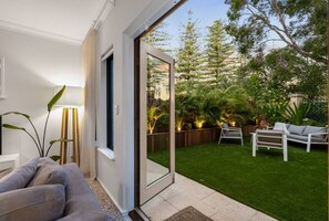 A modern living room with a sofa, a coffee table, and a large sliding glass door leading to a well-maintained backyard with a lawn, patio furniture, and trees. - Cottesloe Shorehouse (Cottesloe)