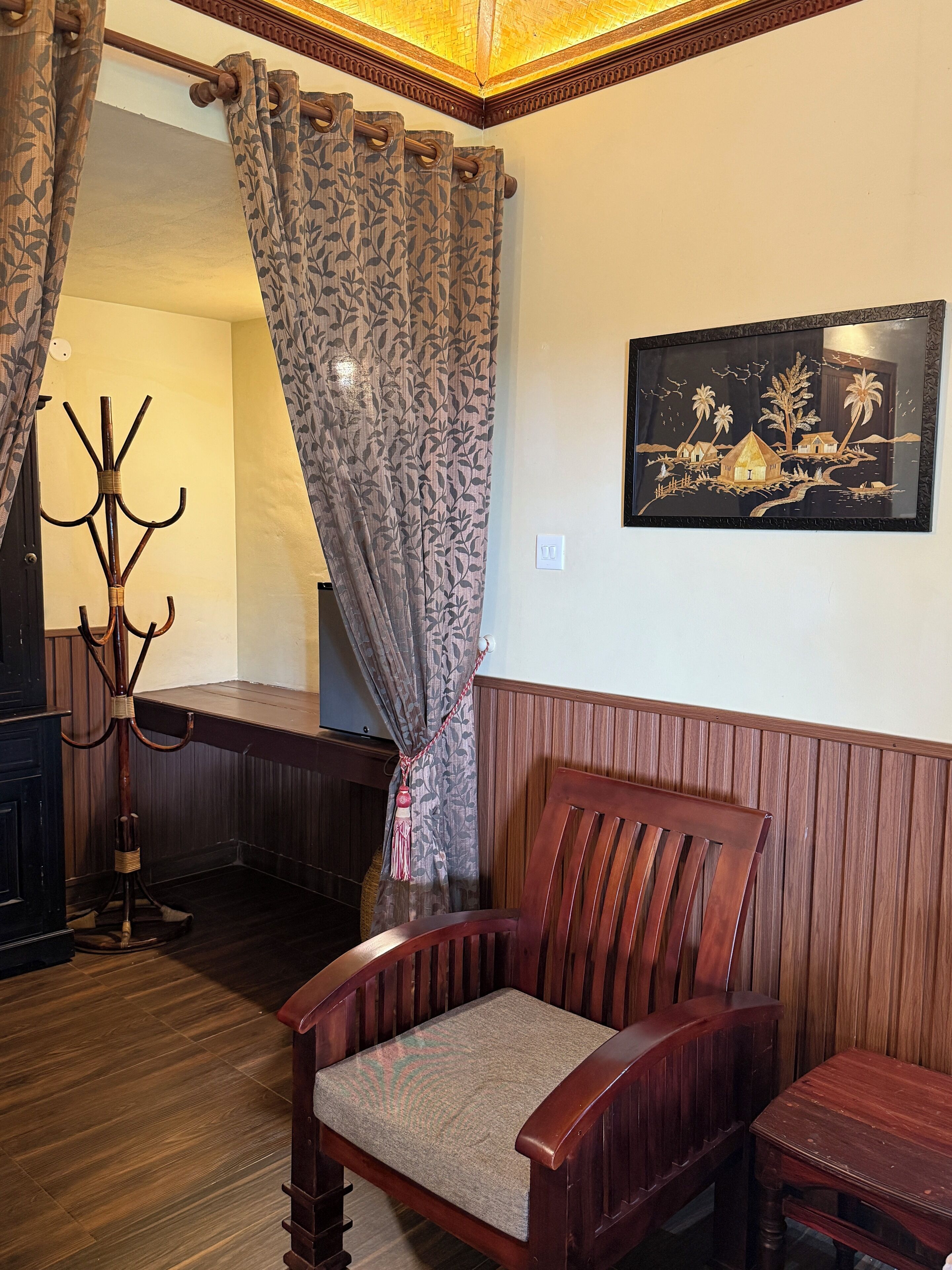 A wooden chair with a cushioned seat, a coat rack, and a painting on the wall.