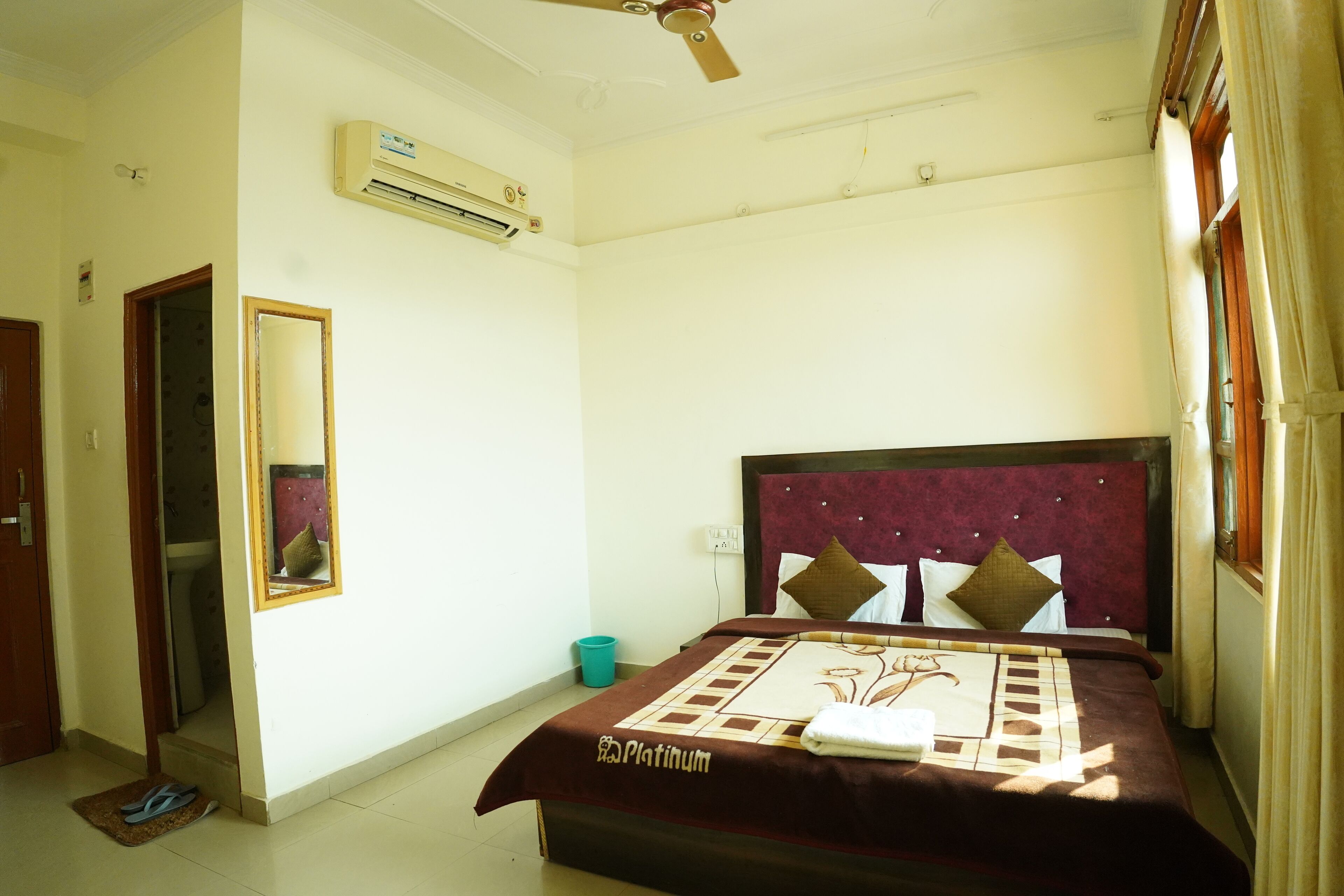 A hotel room with a bed, an air conditioning unit, a ceiling fan, a mirror, and a door.