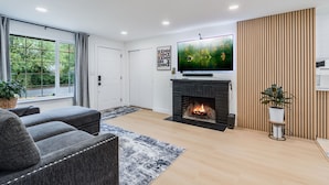 A modern living room with a fireplace, a large flat-screen TV, grey sofas, and a wooden accent wall. - Charming 2-bedroom house in welcoming University Place with AC (University Place)