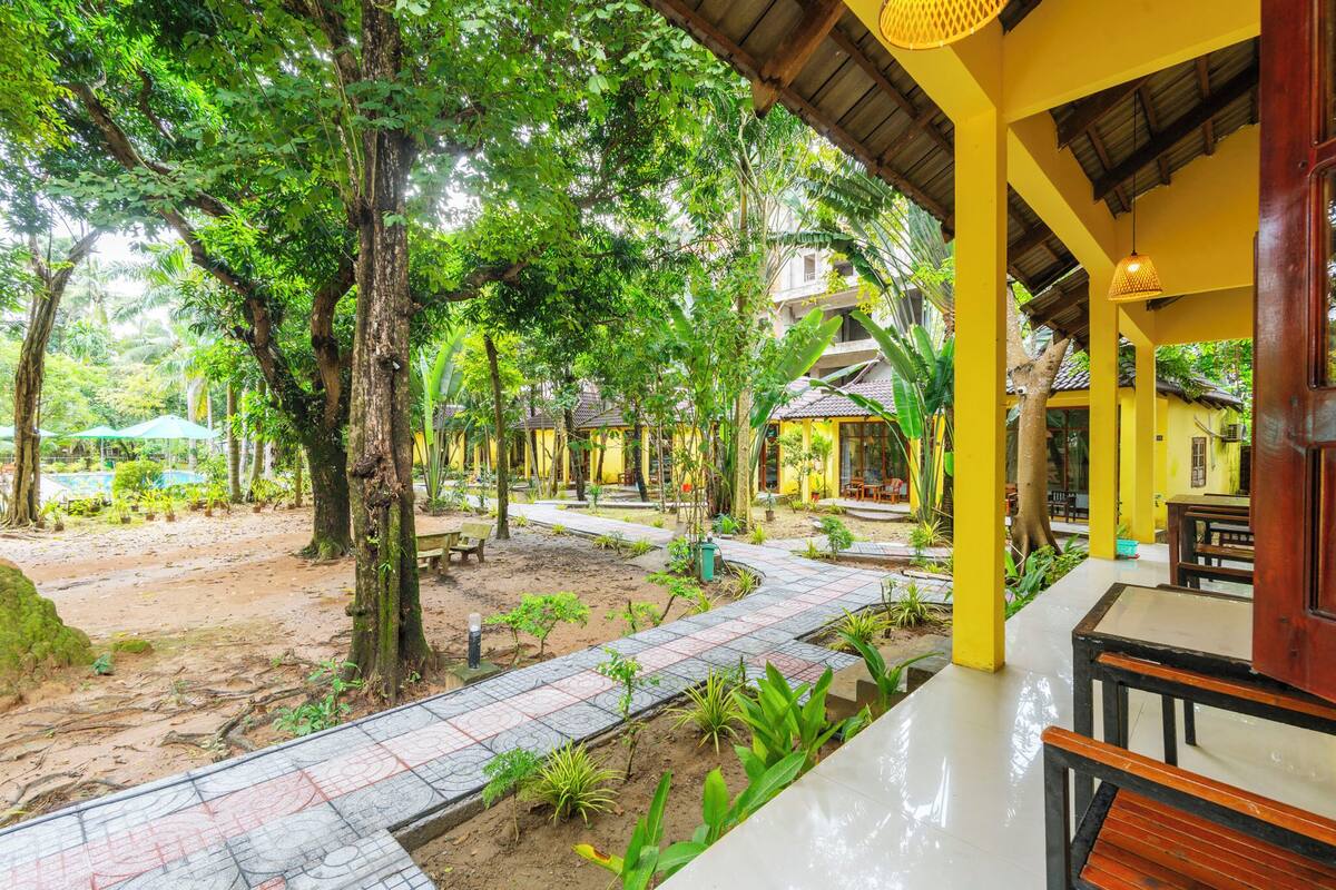 Un patio con un sendero adoquinado, rodeado de árboles y plantas, que conduce a un edificio de fachada amarilla y mobiliario de madera.