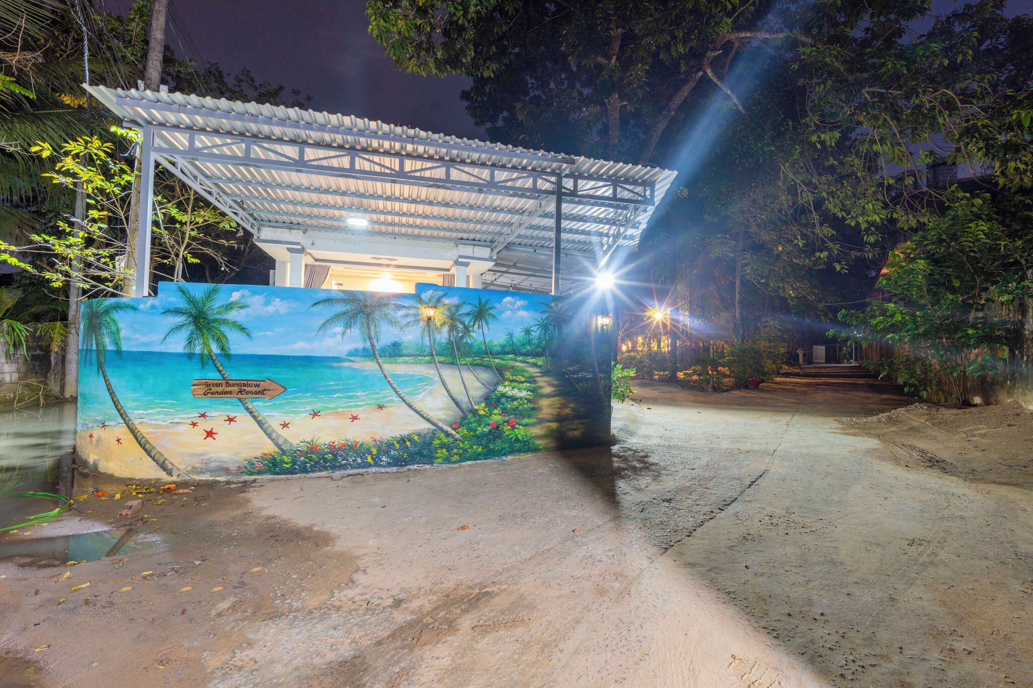 A beach-themed mural on a building's exterior wall, a covered structure, and a concrete pathway leading to the building.