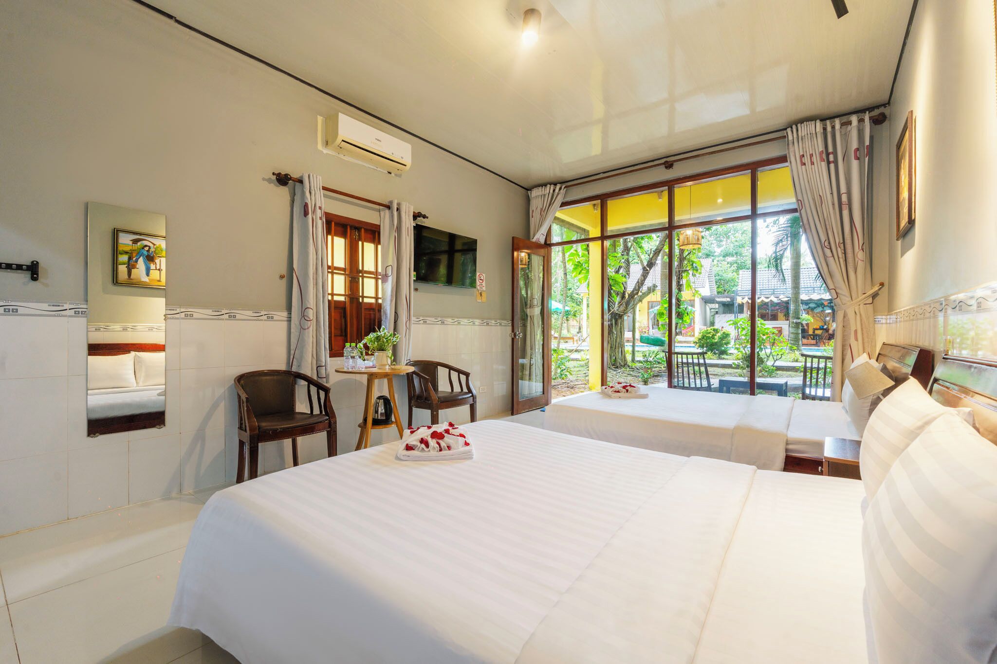 A hotel room with a large bed, a small table with chairs, a window with curtains, and a view of greenery outside.