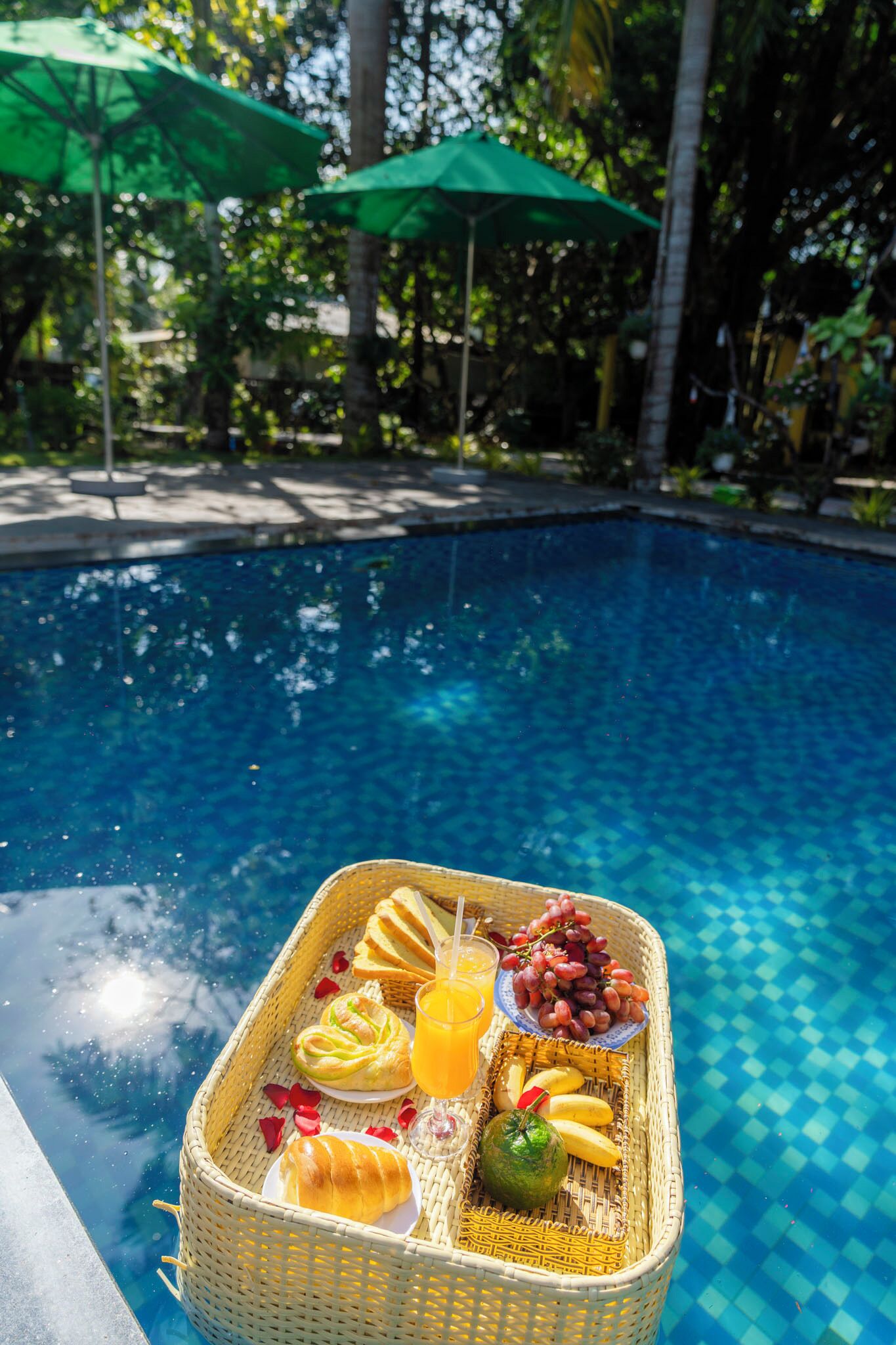 A poolside breakfast setup with a wicker basket containing croissants, bananas, grapes, and orange juice.