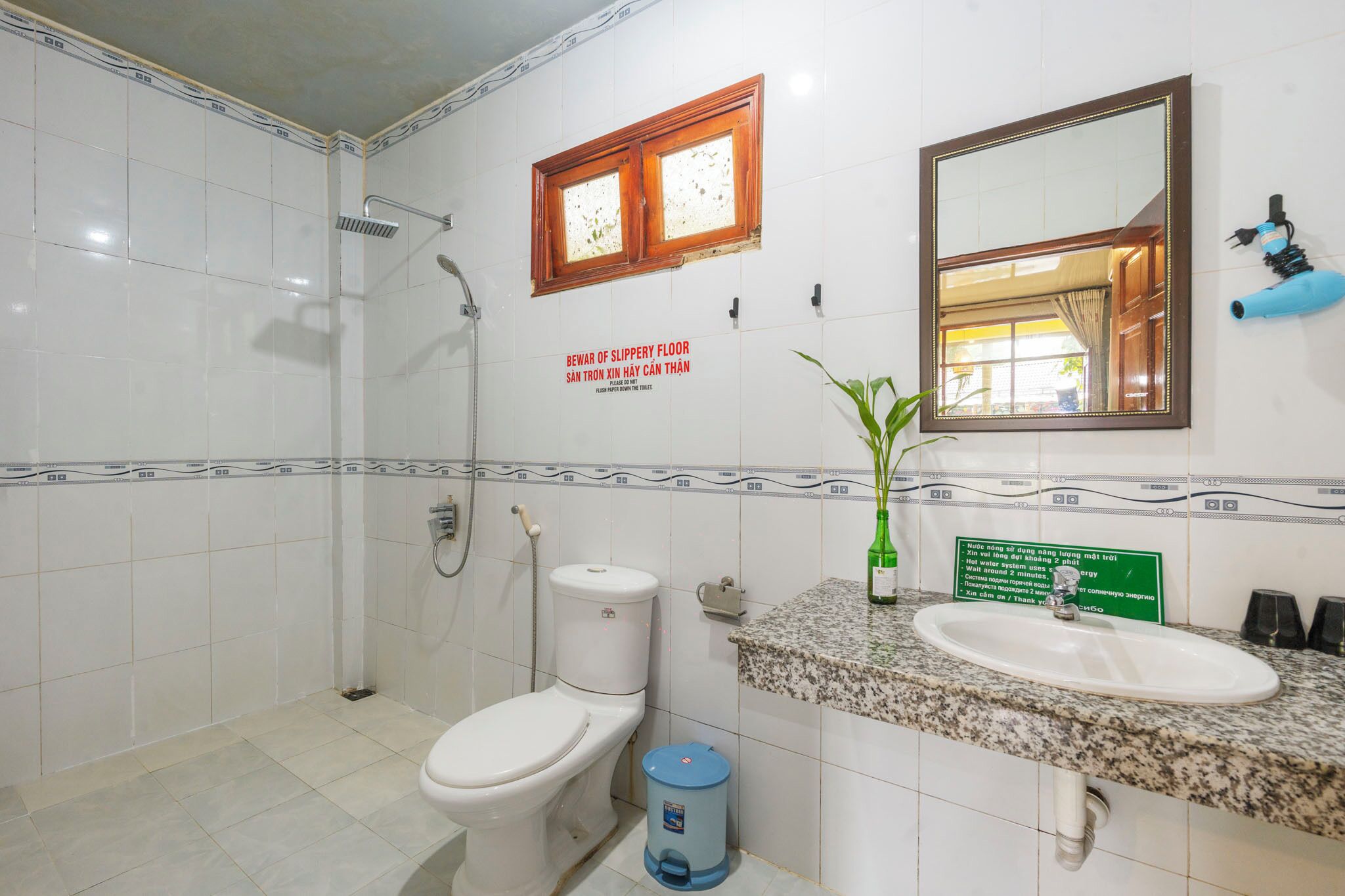 Baño con inodoro, lavamanos y ducha. Hay un cartel que advierte sobre un piso resbaladizo.
