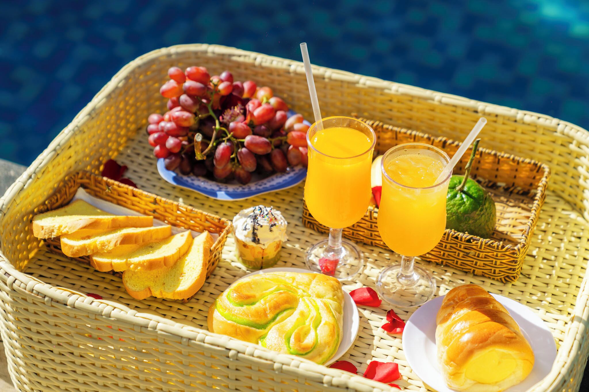 A wicker tray with food and drinks, including grapes, toast, muffins, and croissants, by a pool.