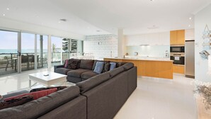 Ein modernes Wohnzimmer mit einem großen, grauen Ecksofa, einem weißen Couchtisch und einem Essbereich mit Glastisch, der auf den Ozean blickt.