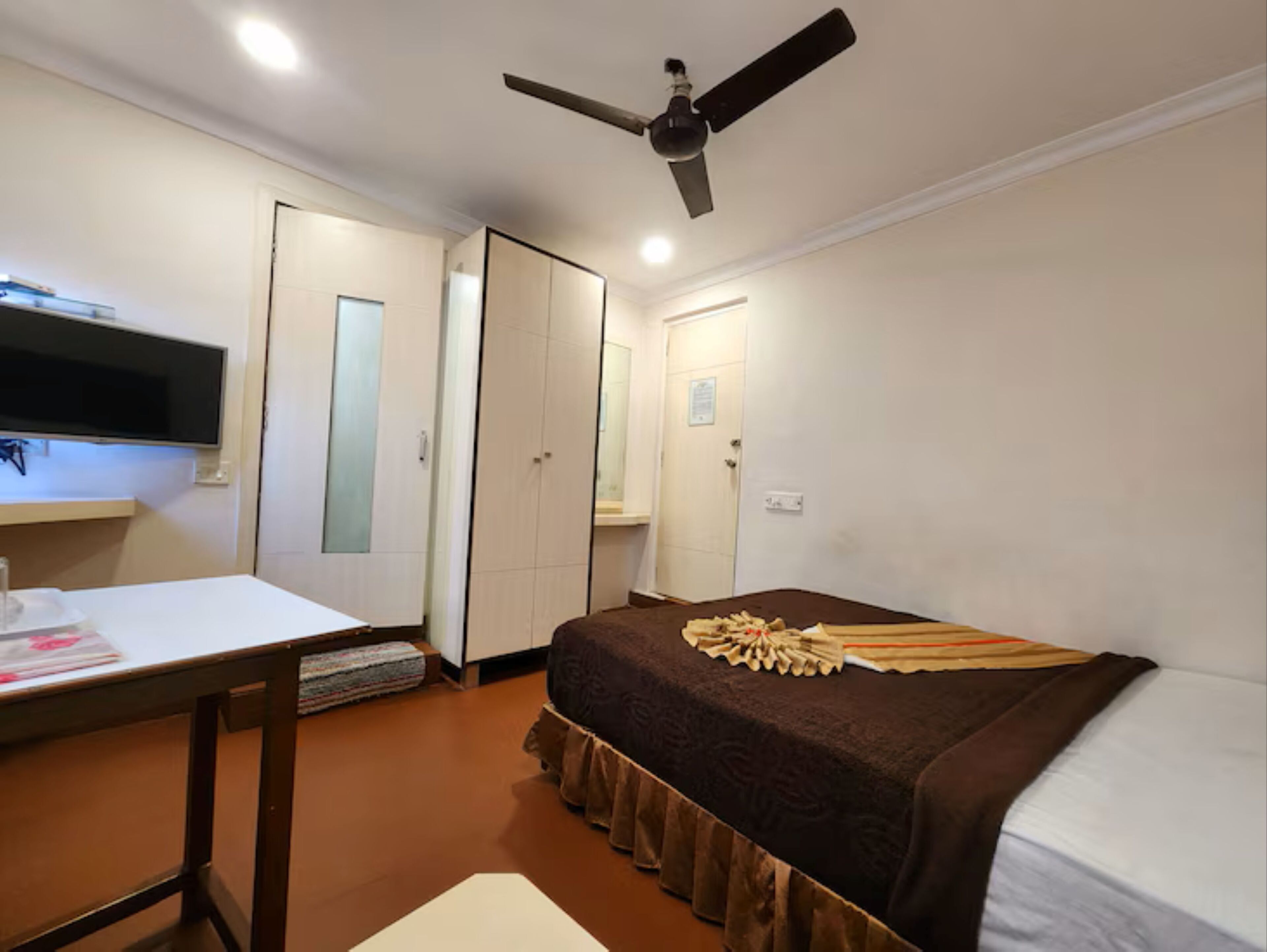 A hotel room with a bed, a desk with a chair, a television mounted on the wall, and a ceiling fan.
