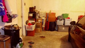 Un garage encombré, rempli de divers objets et de cartons, une voiture garée à droite et une porte à gauche.