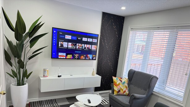 A modern living room with a grey sofa, a white console table, a large flat-screen TV mounted on the wall, a potted plant, and a window with blinds. - Cozy, Private Suite, Two Level Apartment With Separate Entrance (Brampton)