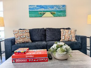 Un salon avec un canapé, deux coussins à motifs, une table basse avec des jeux de société, une télécommande et un vase rempli de fleurs.