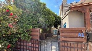 A gated entrance with a wooden gate and a metal fence, a house with a blue door, and a lush green hedge with red flowers. - Newly Renovated Apt with Private Garden - 7 Min Walk to Beach (Paphos)