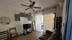 A living room with a brown leather sofa, a TV on a stand, a ceiling fan, and a clock on the wall. - Newly Renovated Apt with Private Garden - 7 Min Walk to Beach (Paphos)