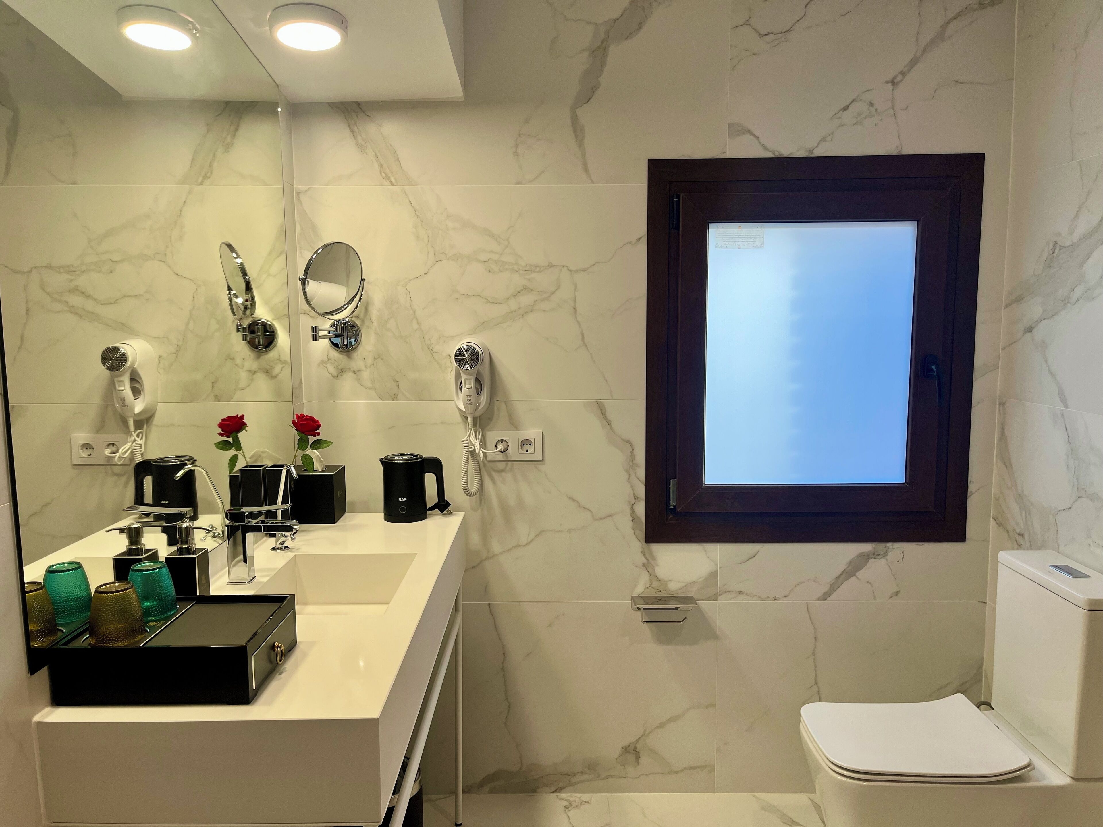 Ein modernes Badezimmer mit Marmorrückwand, einem großen Spiegel, einem Föhn, einer Kaffeemaschine und einer Toilette.
