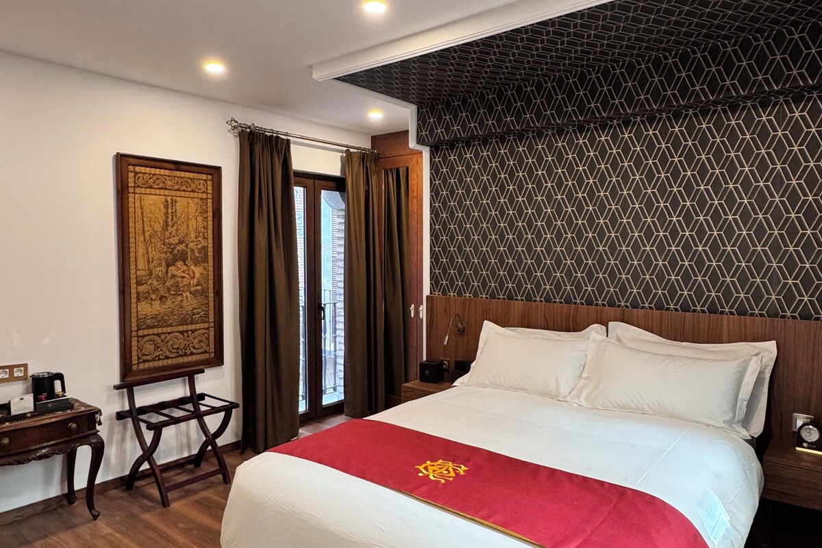 Habitación de hotel con una cama grande, una mesita de noche de madera, un escritorio con una silla, un cuadro en la pared y un papel tapiz estampado.