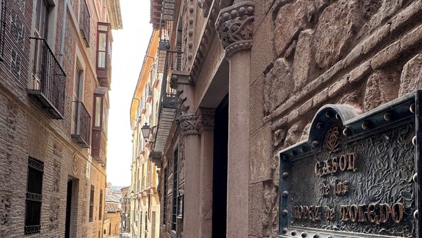 Una estrecha calle adoquinada bordeada por edificios históricos con fachadas ornamentadas y un cartel que indica el lugar como "Cason de la Toledan".