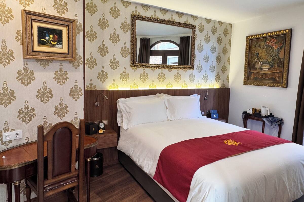 A hotel room with a large bed, a wooden nightstand, a desk with a chair, a mirror, and a framed picture on the wall.