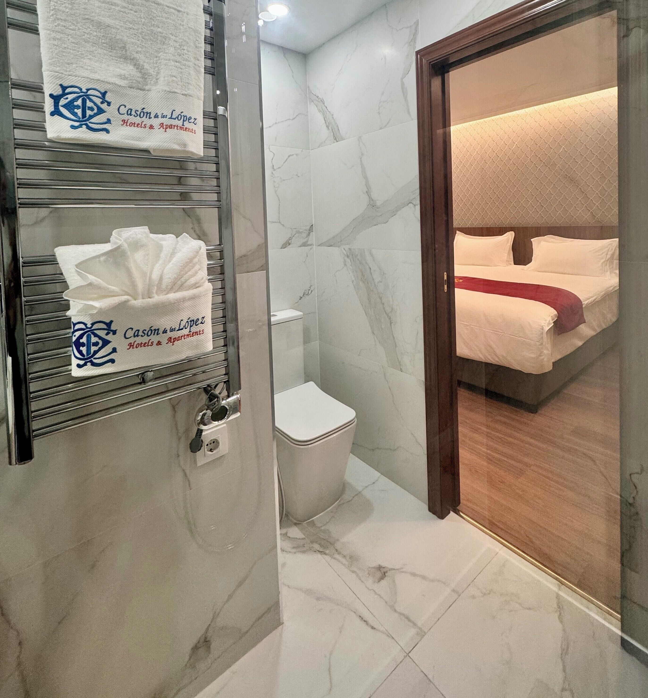 Un baño de hotel con suelo de mármol, un inodoro y una cama visible a través de una puerta de cristal.
