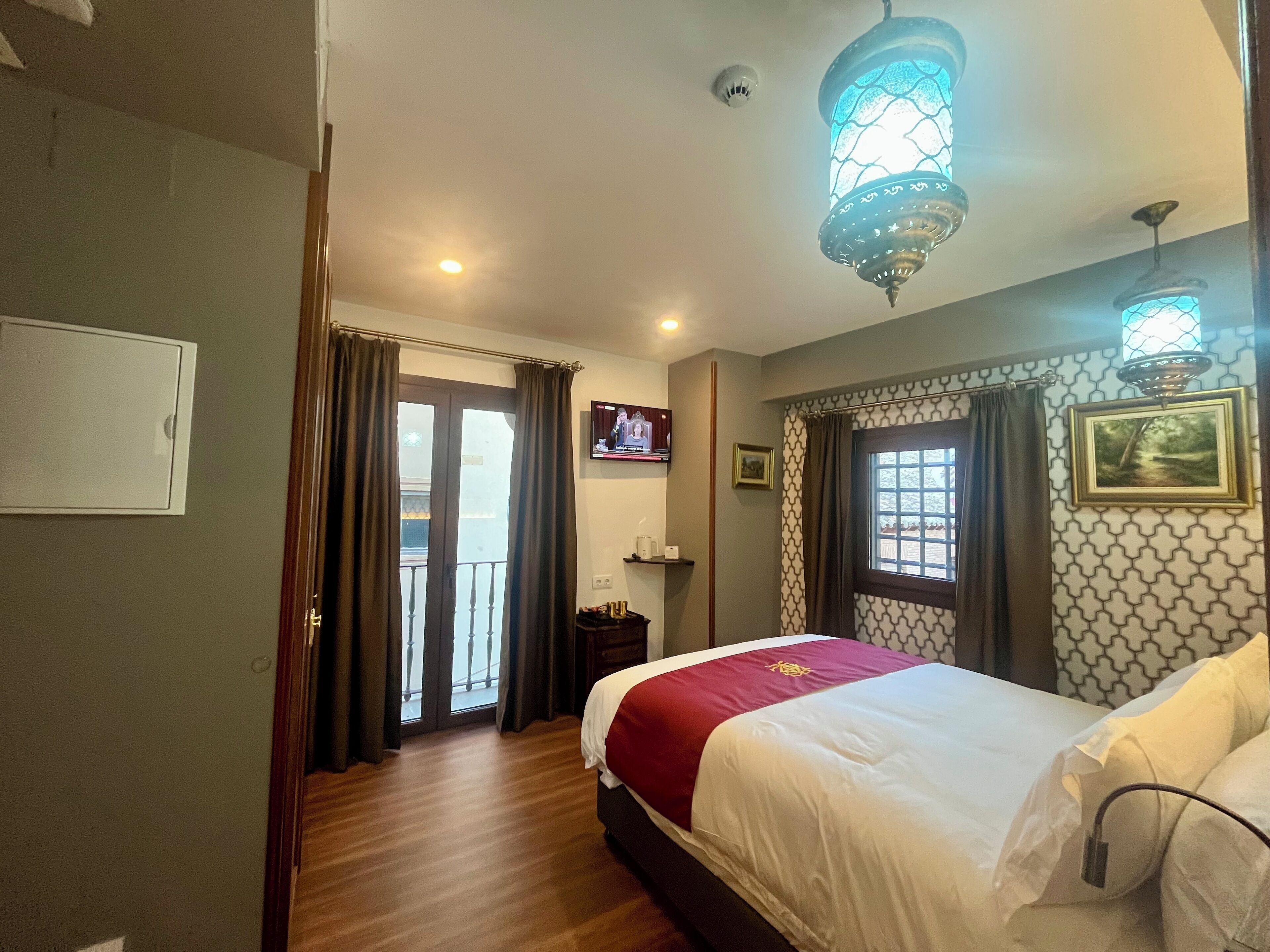 A hotel room with a bed, a television, a window with curtains, and a balcony door.