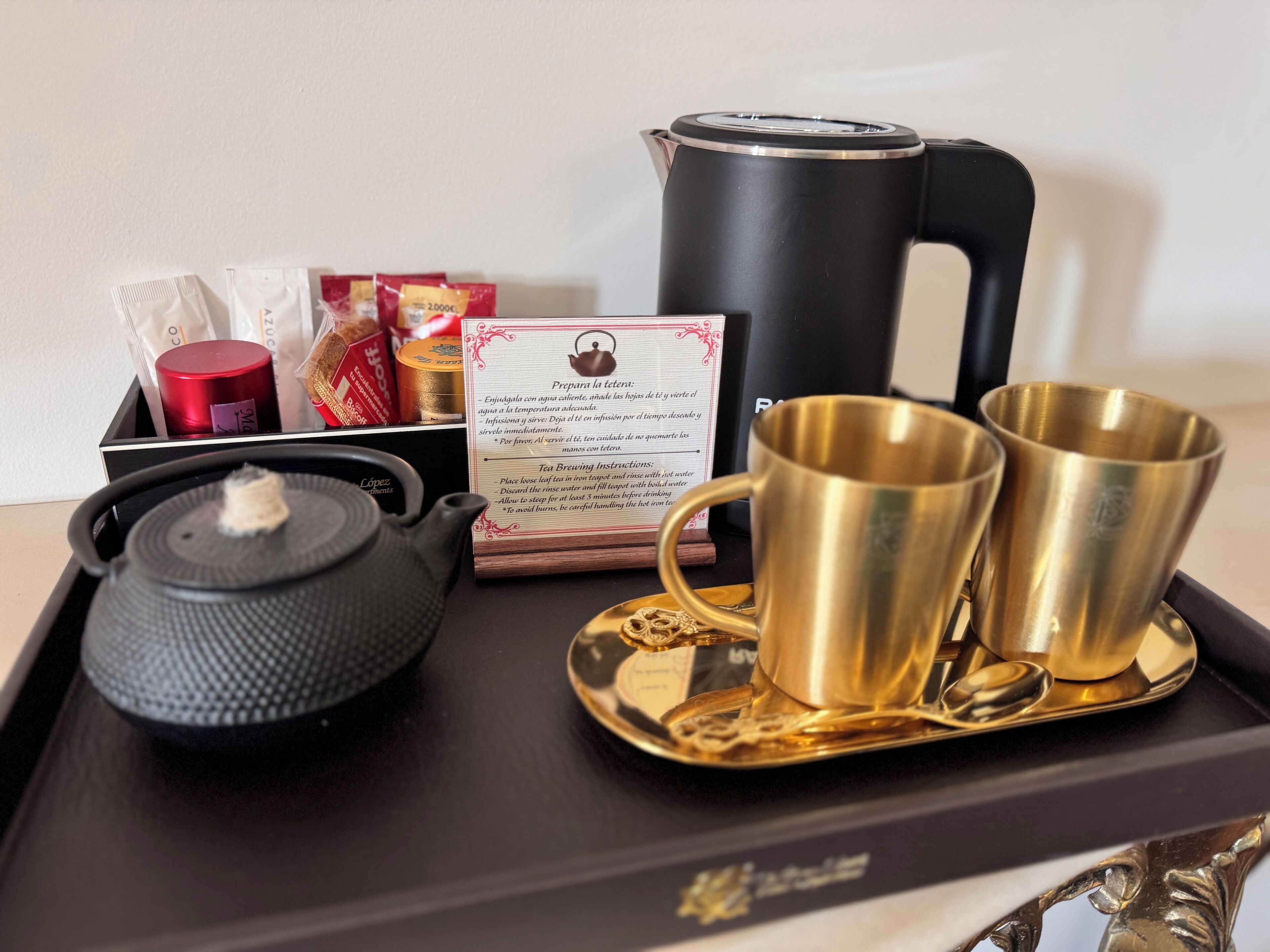 A tray with a black electric kettle, two gold cups, a black teapot, and a tea bag box.