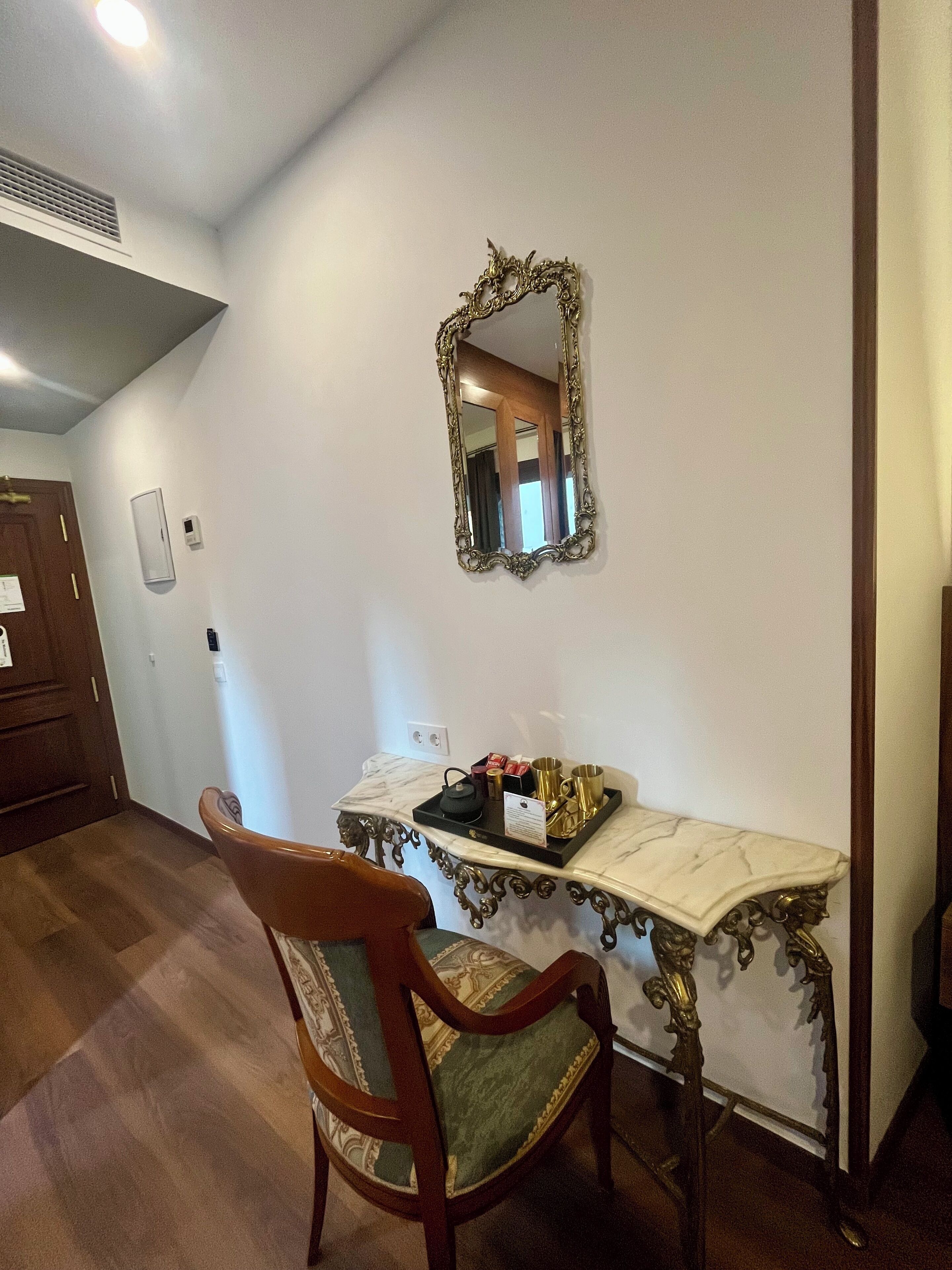A room with a marble console table, a wooden chair with a green cushion, a gold-framed mirror, and a tray with items on the table.