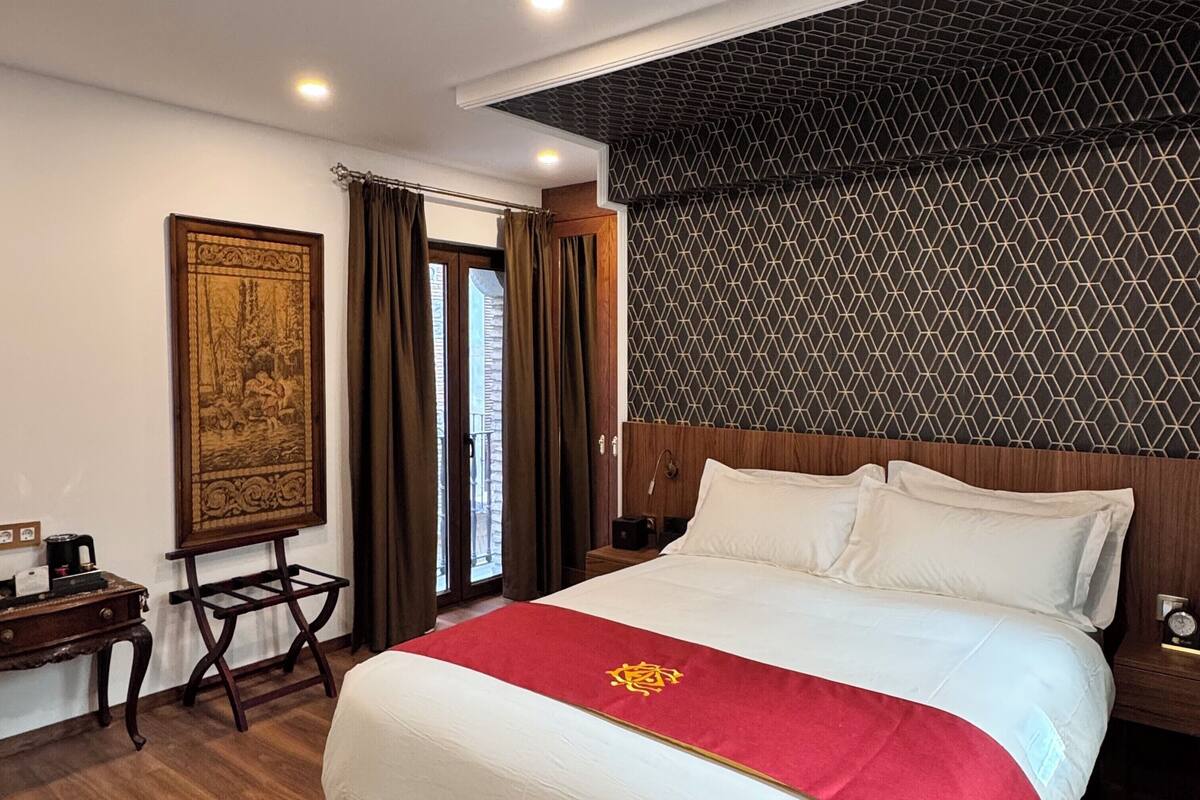A hotel room with a large bed, a wooden nightstand, a desk with a chair, a framed picture on the wall, and a patterned wall.