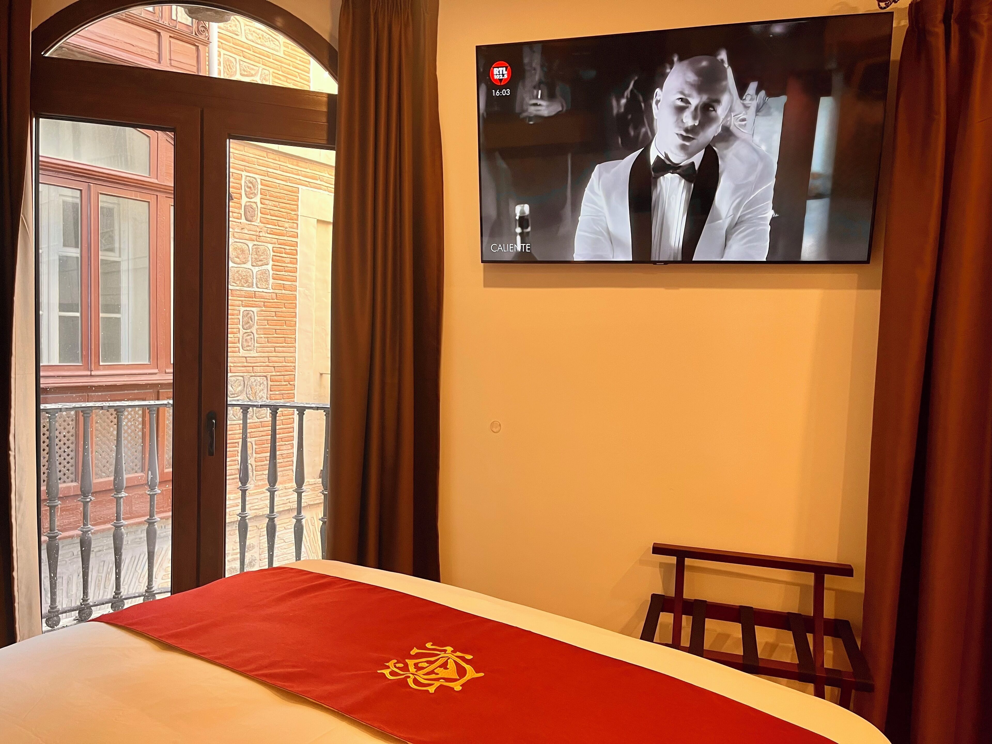 Habitación de hotel con cama, colcha roja con un emblema dorado, televisor montado en la pared y balcón con vista a un edificio de ladrillo.