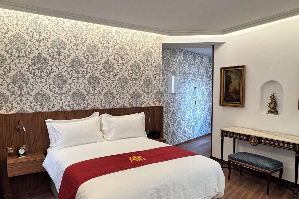 A neatly made bed with a red and white bedspread, a wooden nightstand with a clock, a bench, and a wall with patterned wallpaper and a framed picture.