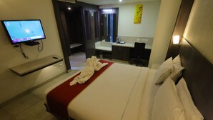 A hotel room with a bed, a TV mounted on the wall, a bathroom with a sink and mirror, and a red carpet. - Breeze Palace (Karur)
