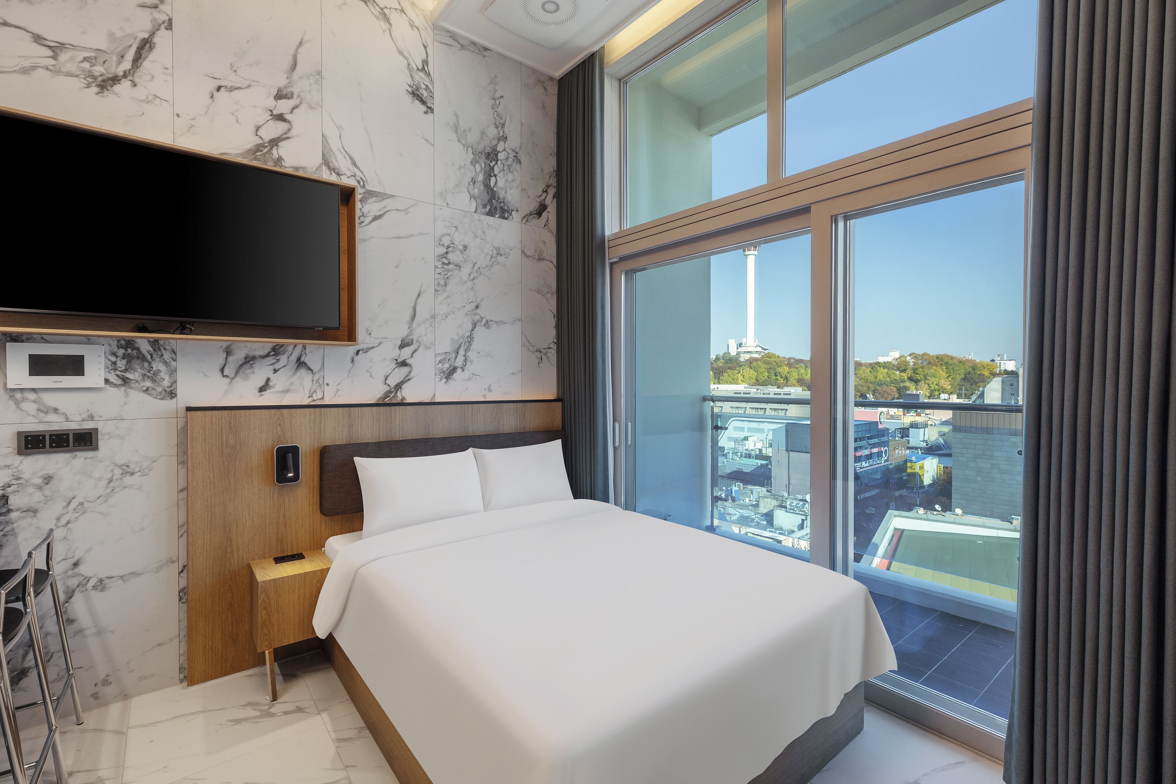 A modern hotel room with a large bed, a TV mounted on the wall, and a view of the city through large windows.