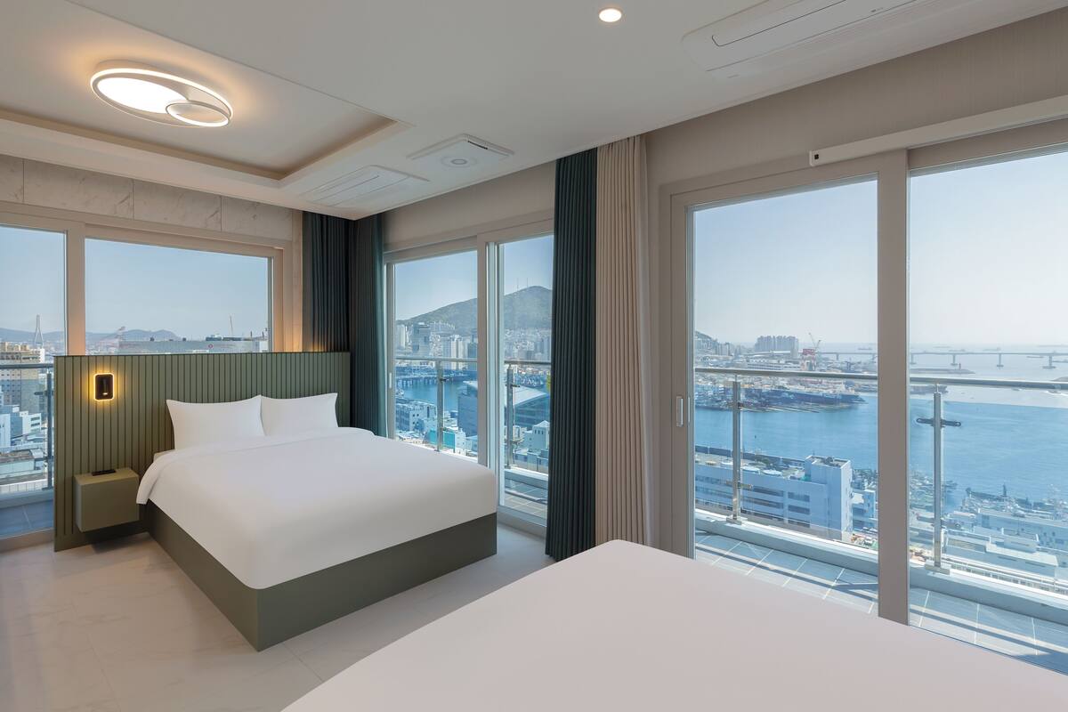 Une chambre d’hôtel moderne dotée d’un grand lit, d’une table de chevet, d’une lampe fixée au mur et de grandes fenêtres offrant une vue sur la ville.