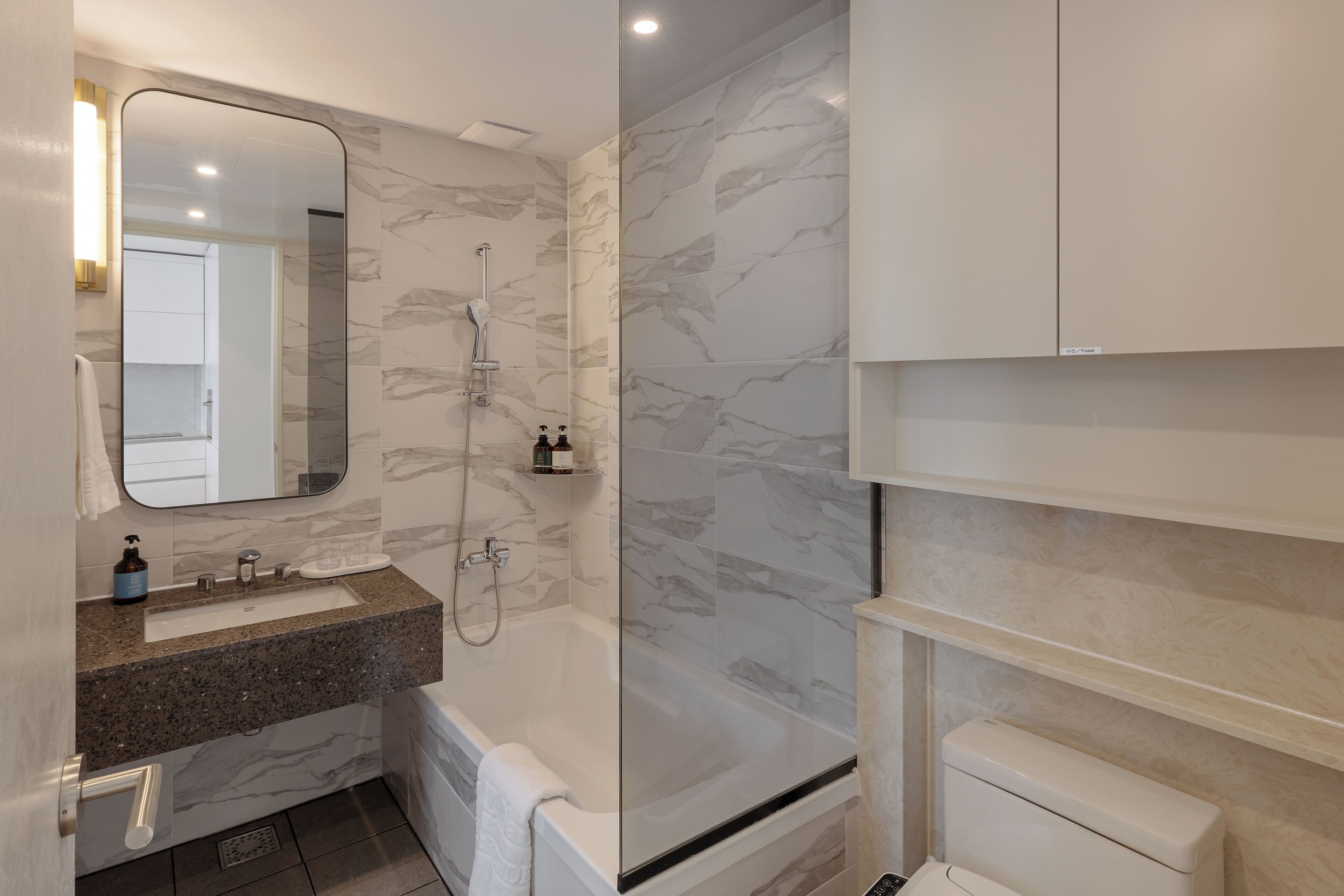 Un baño moderno con pared de mármol, ducha con cerramiento de vidrio, lavamanos con dispensador de jabón y un inodoro.