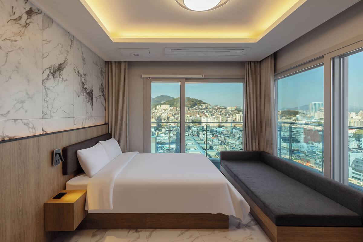 Une chambre d’hôtel moderne dotée d’un grand lit, d’une banquette intégrée et offrant une vue sur la ville grâce à de grandes fenêtres.