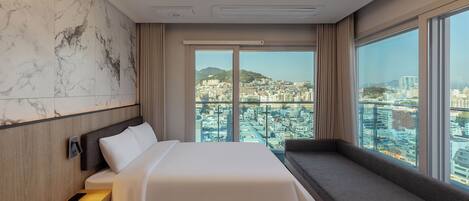 Habitación de hotel moderna con una cama grande, un banco integrado y vistas a la ciudad a través de amplias ventanas.