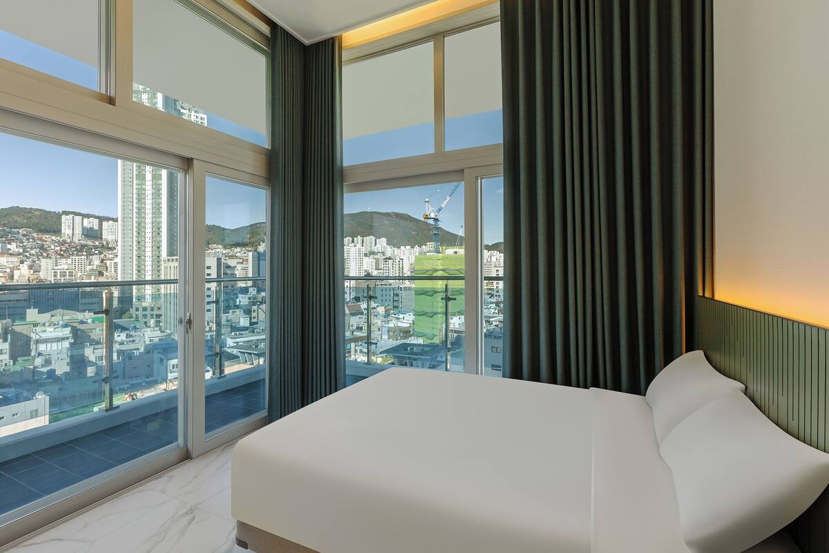 Une chambre d’hôtel moderne dotée d’un grand lit, offrant une vue sur le paysage urbain grâce à de grandes fenêtres du sol au plafond, et disposant d’un balcon.