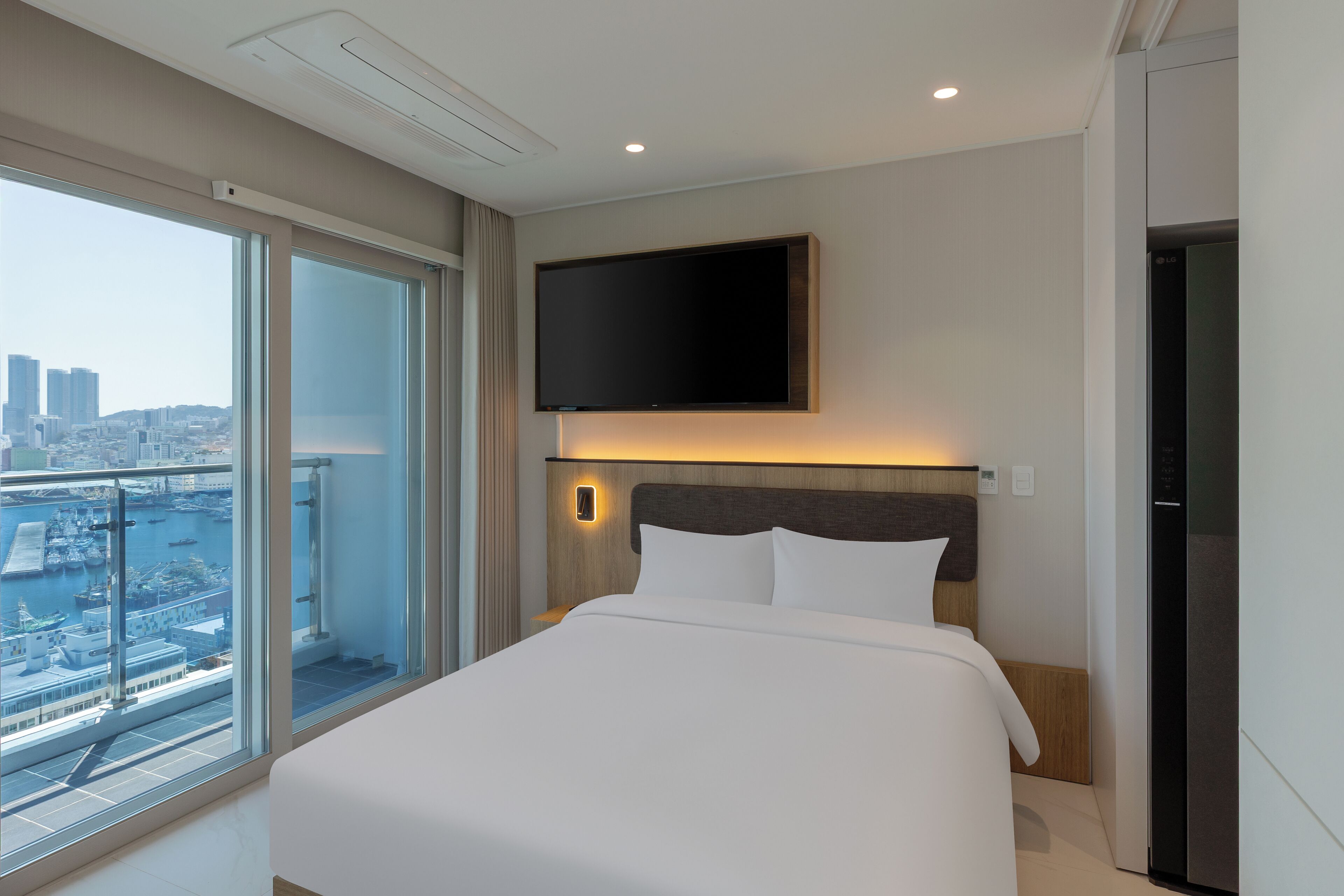 A modern hotel room with a large bed, a mounted TV, and a city view.